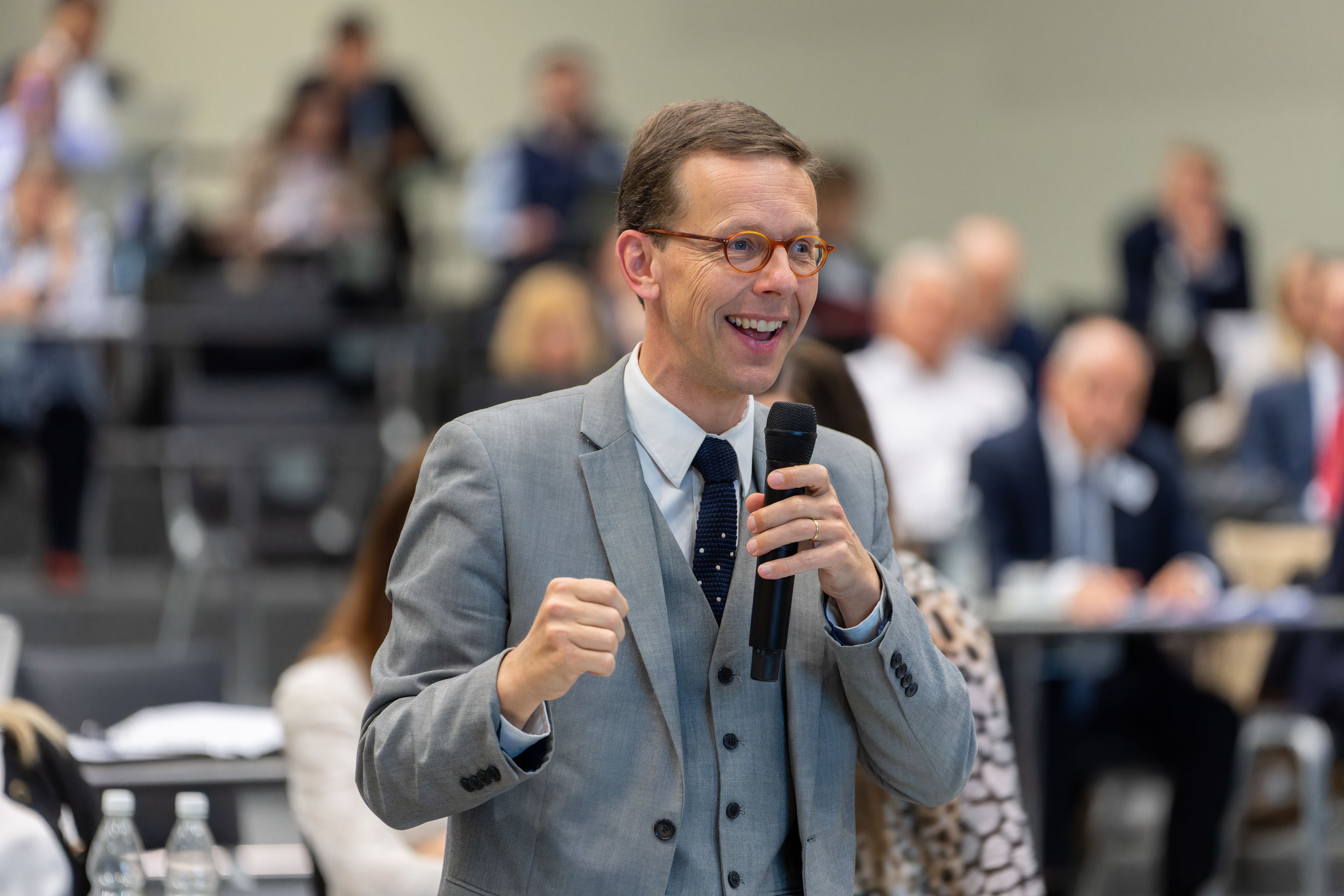 A white man wearing glasses and a grey suit holds a microphone, smiling. 